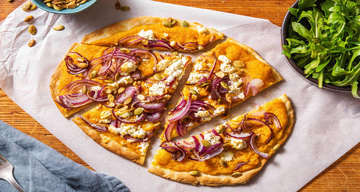 Tarte flambée avec une sauce de potiron à la crème, de la feta et de l'oignon rouge Garnie de graines de potiron et accompagnée de roquette